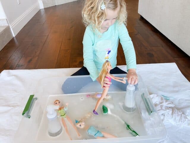 little girl playing with a doll and a soap dispenser in a large plastic storage tub